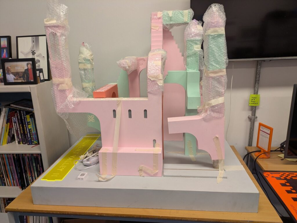 The Monument Valley model in the office at the National Videogame Museum. A number of structures have been moved, secured with masking tape, and wrapped in bubble wrap to protect it during transport. The yellow acrylic sign reading MONUMENT VALLEY II is laid on top of the blue-grey base platform.