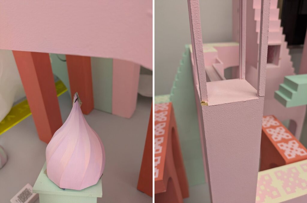 Close-up photo of a tower from the model. One of the four wooden vertical beams has broken at the base and moved slightly out of place. The tower and beams are pink. There are other platforms and stairs visible in the background.
Close-up photograph of part of the model. There is a pink dome with a pointed tip which has cracked at the end. It is sitting loosely on top a light green platform which has some paint worn away, showing the wood underneath. The model's base and several other pillars are visible in the background, and a pink platform runs horizontally across the top of the image.
