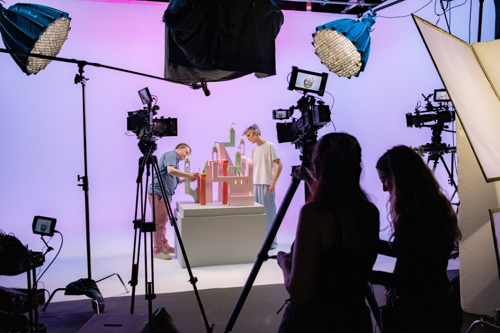Behind the scenes at a photoshoot of an architectural model from the videogame Monument Valley. Two people stand on either side of the model. In the foreground, there are two more people operating lighting and camera equipment. There are multiple lights and camera setups visible.