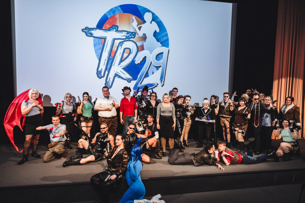 A group around 30 people on a stage, all dressed in Tomb Raider related cosplays.