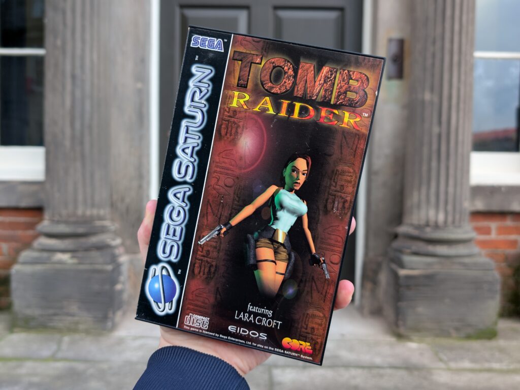 A copy of Tomb Raider for the Sega Saturn. Lara Croft graces the cover wearing a blue tank top, brown shorts, and carries two pistols.