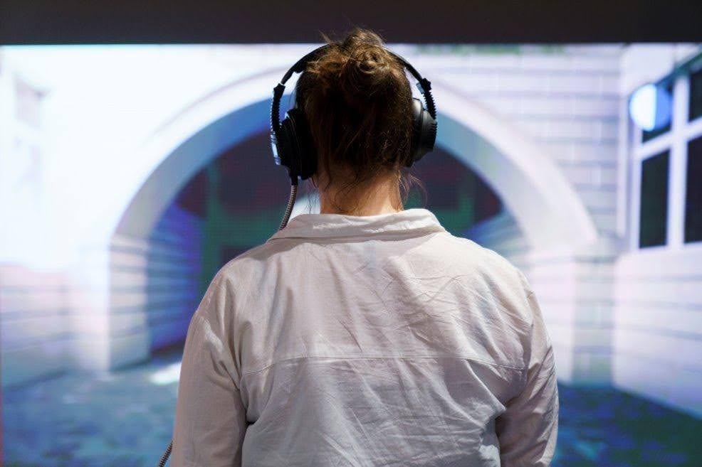 Press photo for the Digital Storytelling exhibition. It shows a person with their back to the camera, staring at a wall with a video being projected onto it. The person is wearing big black headphones connected to something off camera.