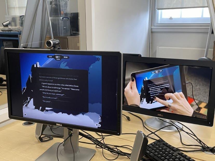 Two screens are shown. One shows a screenshot of a videogame, with a text passage on the screen. The other shows a screenshot of a video of someone using a tablet to play the game, touching the screen with their finger. Both monitors are on a wooden desk.